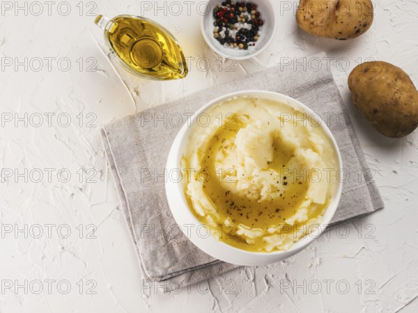 Mashed potatoes in white bowl with cloth napkin, pepper and olive oil on textured concrete white background. Top view or flat lay. Copy space