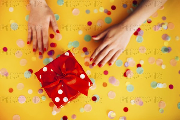 From above of anonymous female with wrapped present decorated with red ribbon and placed on yellow table with glitter and stars