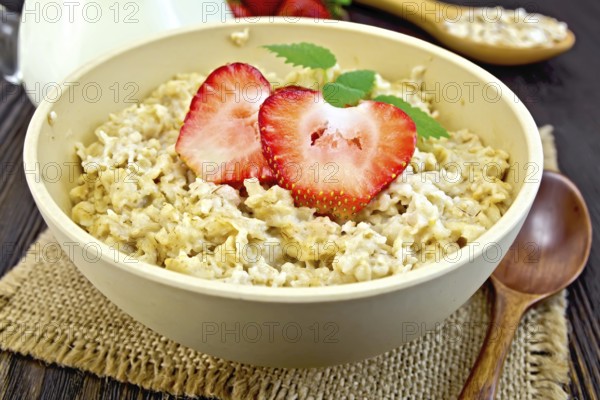 Oatmeal in a yellow cup with strawberries on a napkin of burlap, spoon, milk in a glass jar on the background of dark board