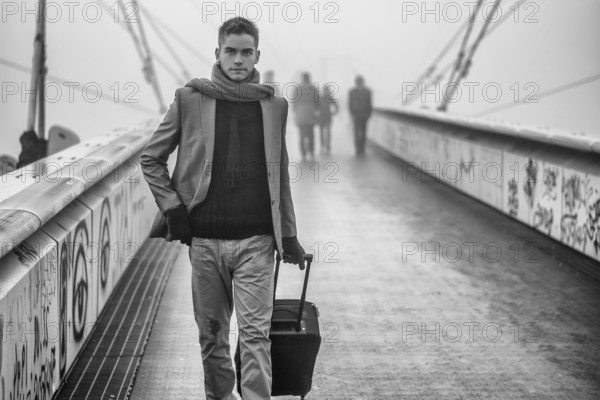 Trendy handsome man walking across a bridge on a misty cold day pulling a suitcase behind him as he departs on a trip
