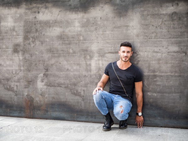 Handsome young man standing against concrete wall, looking at camera, wearing black t-shirt and jeans. Full length shot