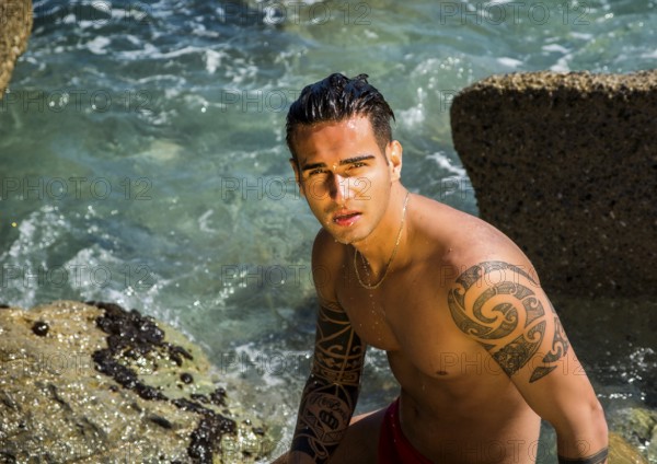 Attractive young shirtless athletic man standing in water by sea or ocean shore, wearing shorts, looking down to a side