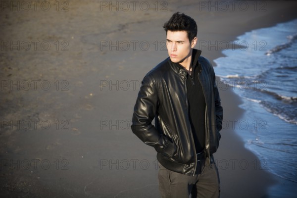 Handsome fashionable young man at the seaside on the beach with his black leather jacket looking away, large copyspace