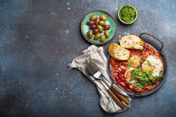 Middle Eastern and Maghrebi healthy dish Shakshouka made of eggs and tomato sauce served in pan with toasts, fresh cilantro and olives on rustic concrete background table from above, space for text, food photography