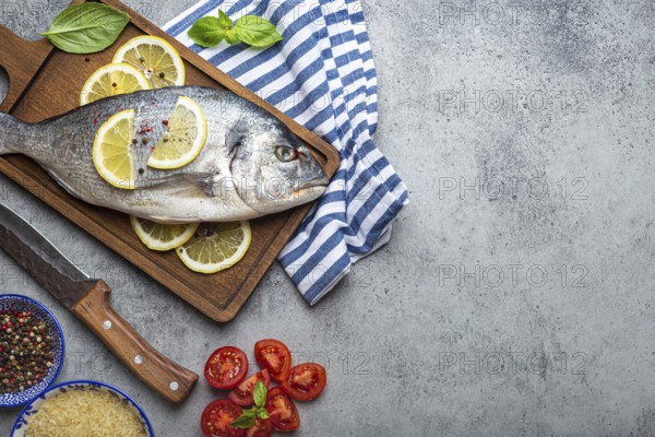 Raw fish dorado with ingredients lemon, fresh basil, cut cherry tomatoes, uncooked rice on wooden cutting board with knife on rustic stone background top view, cooking healthy fish dorado. Copy space, food photography, food photography