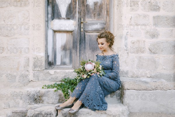 A bride in a stylish blue dress with a bouquet in her hands sits on the steps near an old wooden door .