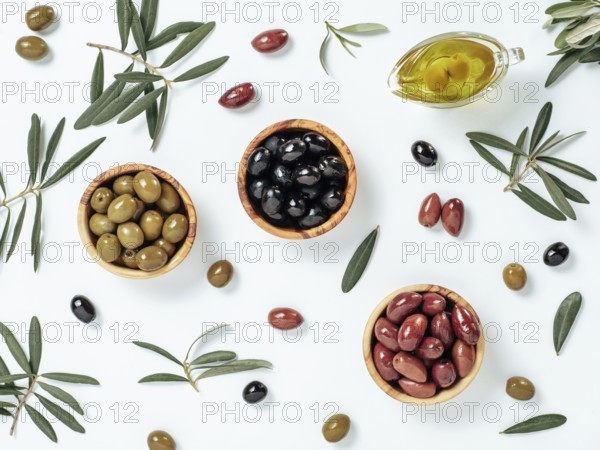 Set of green olives, black olives and red kalmata olives and extra virgin olive oil on white background. Top view of different types of olives in bowls and oil on leaves and branches background
