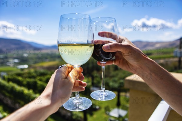 Happy couple hands cheering red and white wine glasses from a winery tasting room terrace against green vineyards landscape, Okanagan Valley BC Canada