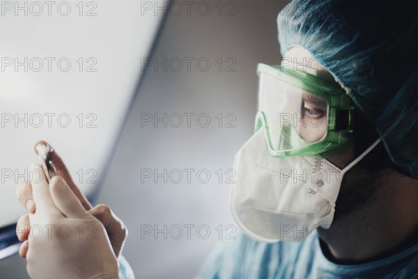 A doctor in green goggles examines a vial while wearing a mask