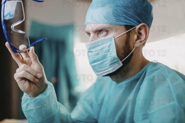 A surgeon attentively holds his protective glasses, wearing a mask