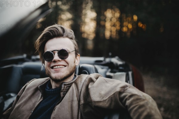 Man smiling broadly in a convertible, wearing sunglasses near a forest at sunset