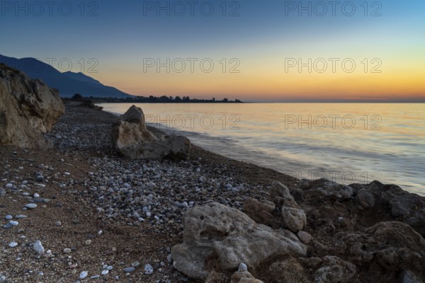 A peaceful sunset over the ocean with silhouette of mountains and pebble beach iwth rock boulders in the foreground