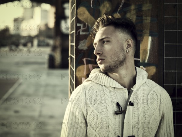 Handsome young man in city setting, with industrial site around him, looking away, serious expression