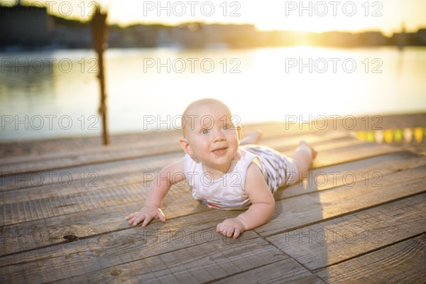 A child a boy, one year old, blond man lies on his stomach on wooden dock, pier in striped clothes, compound near pond on sandy beach against background of river in the summer at sunset of the day
