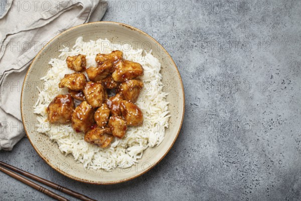Chinese sweet and sour sticky chicken with sesame seeds and rice on ceramic plate with chopsticks top view on gray rustic stone background, traditional dish of China. Space for text, food photography, food photography