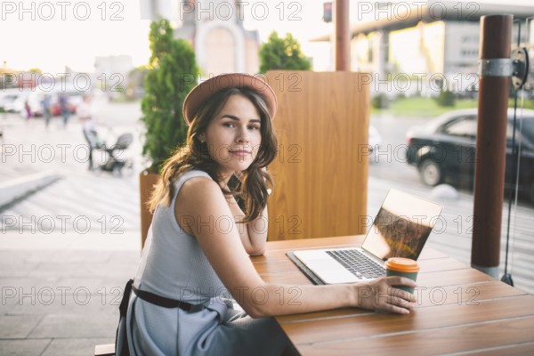 Beautiful young caucasian woman sitting on a terrace in a cafe in summer at a wooden table in a hat and a plate uses technology, working behind a laptop booking hotels trip in the summer