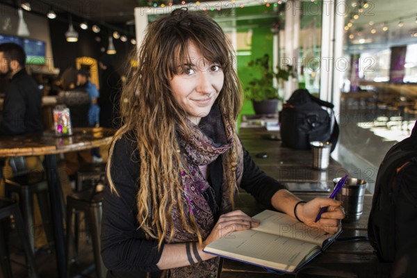 Portrait of smiling beautiful girl with dreadlocks and tattoos. Writing on a notebook with a pen in her hand. Selective focus with blurred background