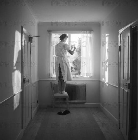 In the 1950s. A woman is seen at a window in a corridor standing barefoot on a chair to hang the window curtains up. 1955. Svahn