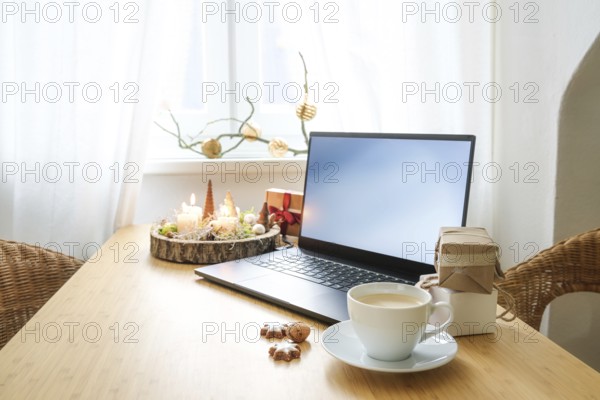 Laptop on a wooden desk by the window in an attic apartment. Gifts, advent candles, Christmas decoration and a coffee cup. Holiday online shopping, copy space, selected focus, narrow depth of field