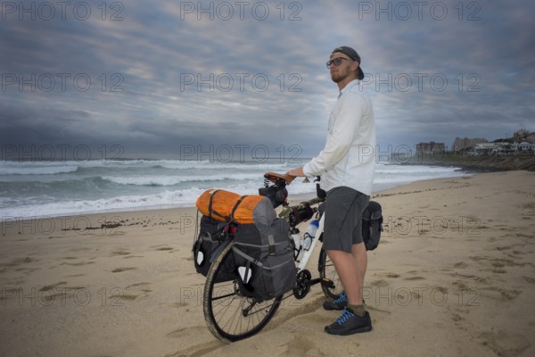 A long distance adventure cyclist stands with his bicycle in hands on the beach while looking at the ocean