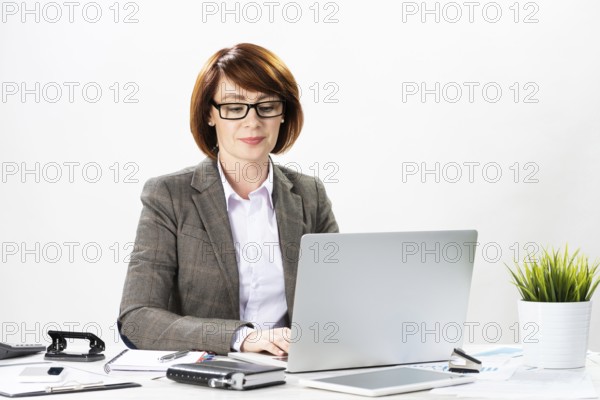 Attractive middle aged businesswoman working with computer in office. Business lady sitting at desk on white background. Successful corporate woman in suit and glasses posing at her workplace