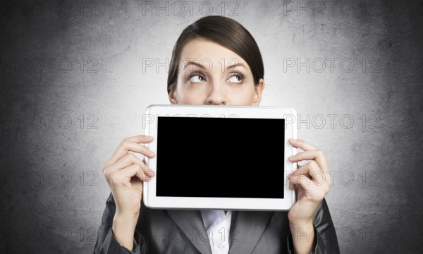 Businesswoman holding tablet computer with blank screen. Beautiful woman in business suit show tablet PC near her face. Corporate businessperson on grey wall background. Digital technology layout