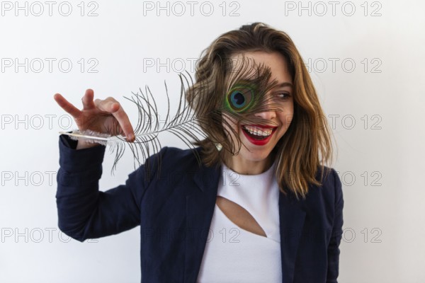 A close up and front portrait of a flirtatious brown haired woman standing against a white background, holding a peafowl tail over one eye and laughing