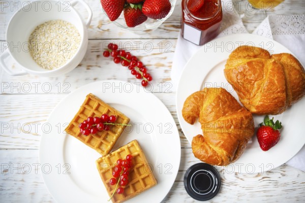 Waffles with red currant jam and berries on a white plate, croissants, orange juice and oat flakes oatmeal on the wooden background