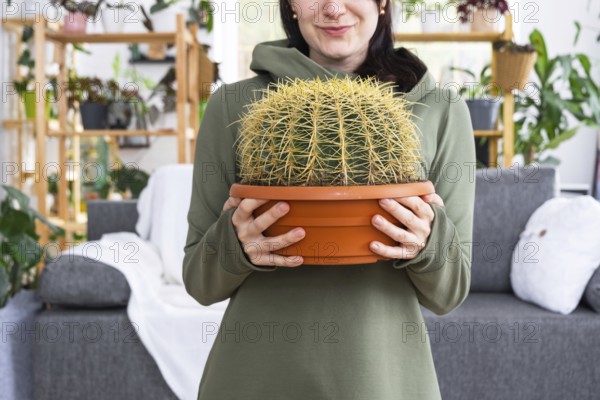 Large echinocactus Gruzoni in the hands of a woman in the interior of a green house with shelving collections of domestic plants. Home crop production, plant breeder admiring a cactus in a pot