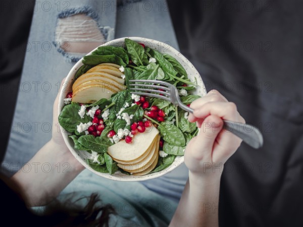 Woman in jeans holding vegan salad bowl with spinach, pear, pomegranate, cheese. Vegan breakfast, vegetarian food, diet concept. Girl in jeans holding fork with knees and hands visible
