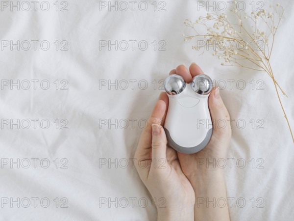 Microcurrent concept. Face massager with micro currents for home use and skincare on white bed. Female hands holds device for anti-aging and face lifting. Top view or flat lay, copy space for text