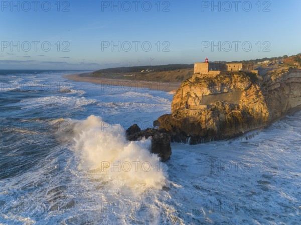 Aerial view of lighthouse on a cliff with a fortress on the coast of the Atlantic ocean with big waves at sunset in Nazare, Portugal, Europe