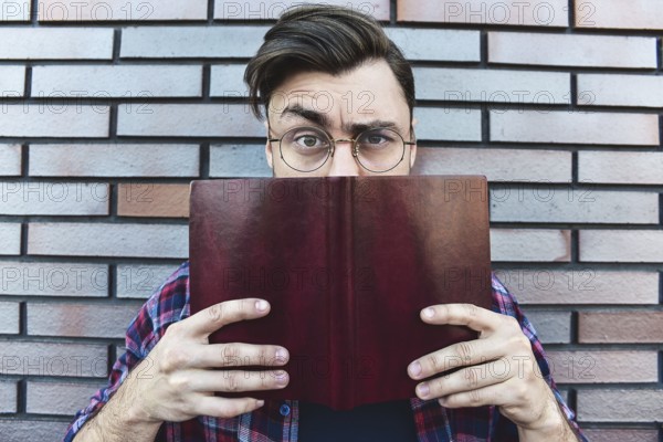 Funny face. Young hipster guy wearing eyeglasses with picked up one browe looking at camera and holding a book or note book on brick wall background