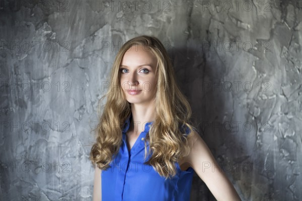 Portrait of the blonde woman in blue blouse staying in front of dark wall and keep smiling