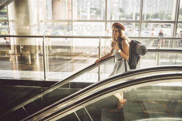 Portrait Smiling Woman Wearing Hat In Airport At Escalator. people travelling with hand luggage. Theme tourism and transport. Caucasian girl with coffee in airport terminal on an escalator