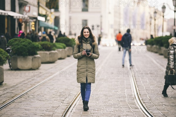 Beautiful young woman tourist with long hair stands on the background of the central street in the city of Munich in Germany in winter. Holds a black big professional camera, poses and smiles