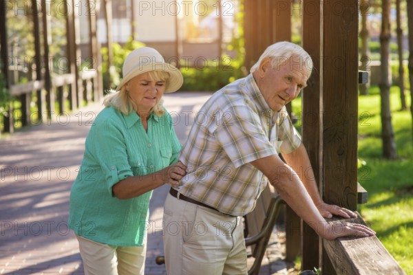 Senior male leans on fence. Elderly couple outdoors. Pains in lower back. Old traumas influence health