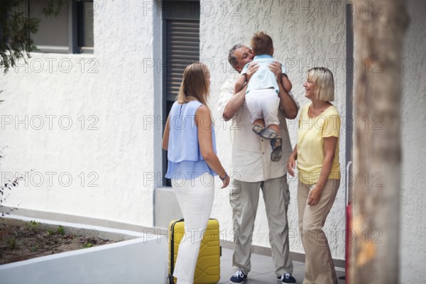Grandfather greeting his young grandchild lifting him in the air and kissing him as he arrives on a trip with his mother outside the apartment