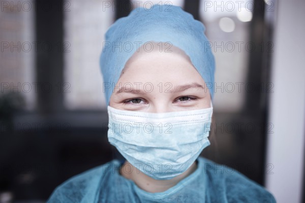 A person in blue scrubs and a medical mask smiles indoors, indicating a healthcare setting