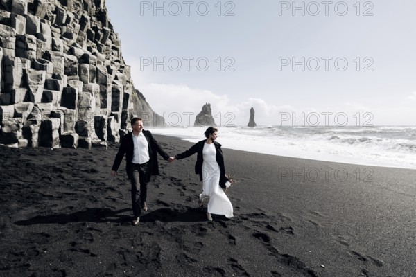 Destination Iceland wedding. A wedding couple walks along the sandy black beach of Vik, near the basalt rock, in the form of pillars. The bride and groom hold hands and walk along the shore