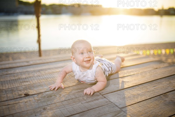 A child a boy, one year old, blond man lies on his stomach on wooden dock, pier in striped clothes, compound near pond on sandy beach against background of river in the summer at sunset of the day