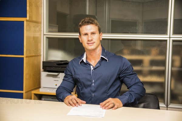 Young man standing, working in an office at the desk with a sheet of paper in front of him, looking at camera
