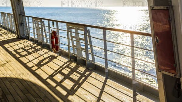 Empty wooden deck and railing with a life ring hanging on the rail on a passenger boat or cruise ship, with sparkling sunshine reflecting on the water of the ocean in a travel concept