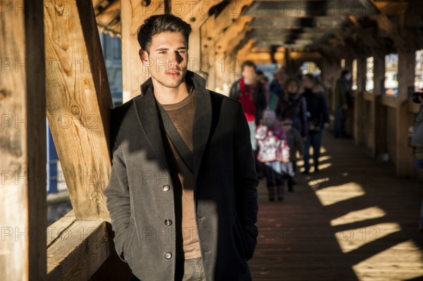 Young dark haired man standing on wooden medieval footbridge in Switzerland while looking away
