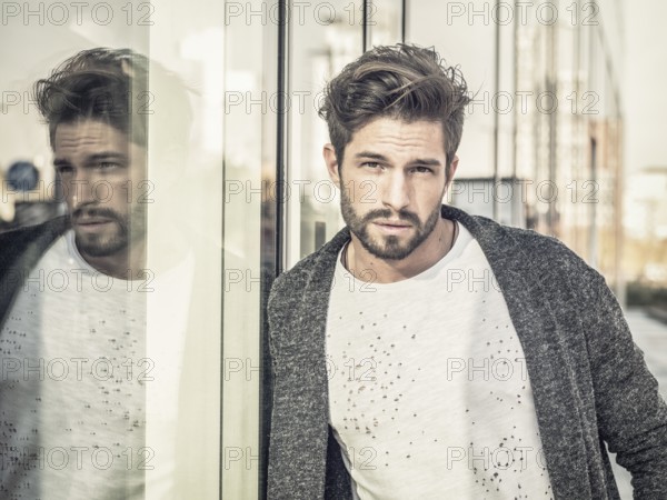 One attractive man in urban environment in city, leaning on glass shop window, wearing cardigan, looking at camera