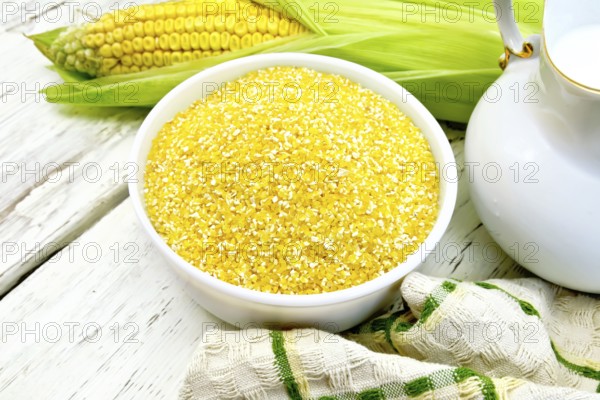 Corn grits in a white bowl, cobs, a jug of milk and a napkin on the background light wooden boards