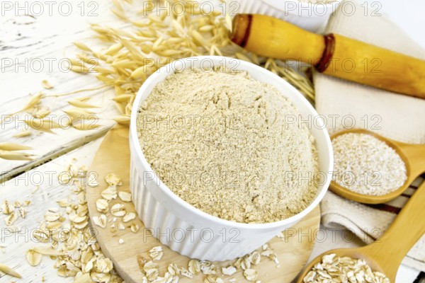 Oat flour in white bowl, oatmeal and bran in wooden spoon, stalks of oats, a rolling pin and a napkin on a wooden boards background