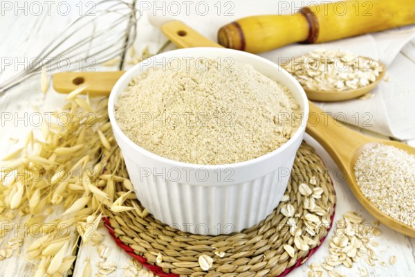 Oat flour in white bowl, oatmeal and bran in wooden spoon, stalks of oats on the background of wooden boards