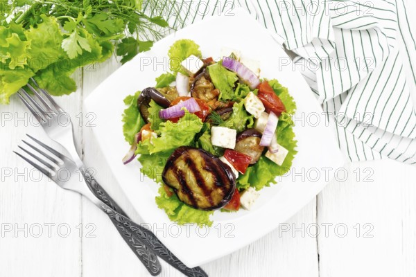 Salad of fried eggplant, fresh tomato with red onion and salted feta cheese, seasoned with vegetable oil and soy sauce on lettuce in plate, napkin and parsley on wooden board background from above