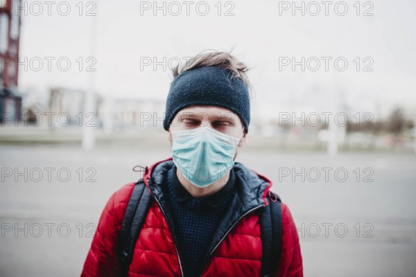 Man in a red jacket and face mask outdoors, with city buildings in the background
